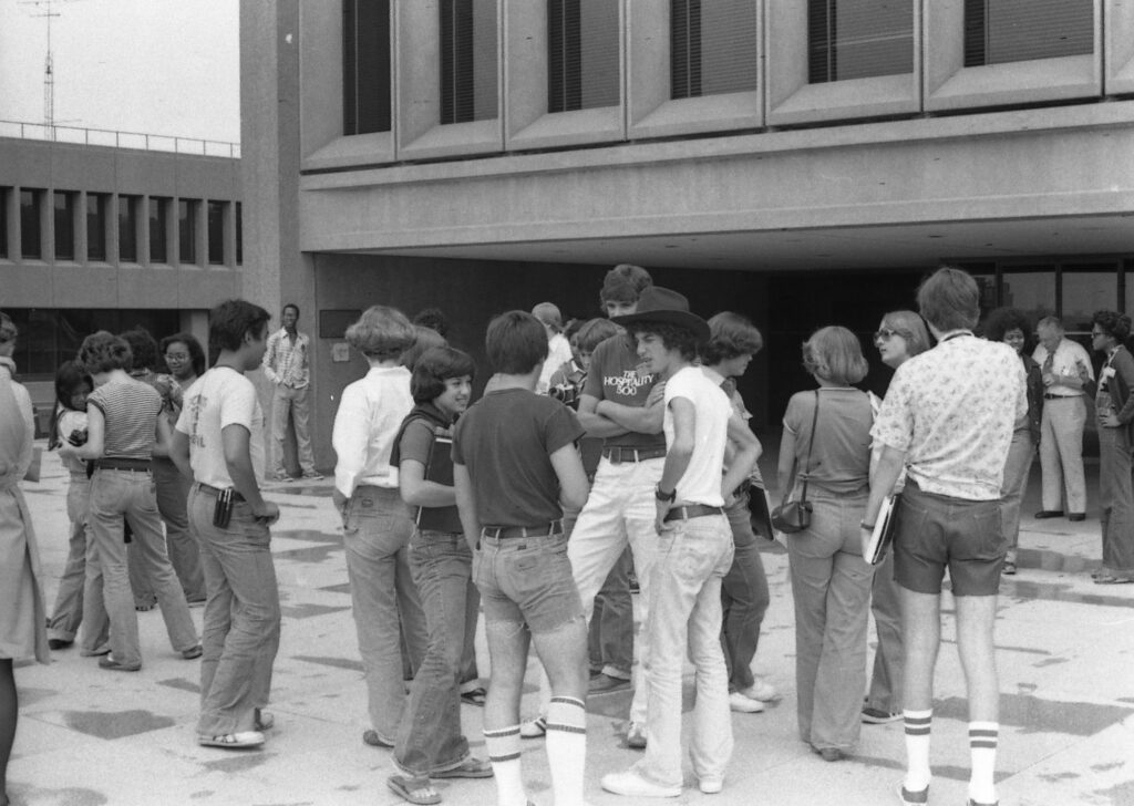 Photograph showing a large group of high school students standing around on a composite stone plaza outside I.M. Pei's Newhouse School at Syracuse University. They are mostly wearing denim jeans and t-shirts of various types. Some of the boys have shorts and stripe-topped tube socks. The jeans are bell-bottomed or at least flared, the belts are wide. The girls are dressed about the same as the boys.