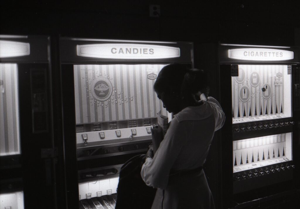 1977 black and white photograph. In a darkened room, nearly all the illumination appears to come from a row of vending machines – to the left can only be seen partially, but in the center is one labeled "candies," and the next one is labelled "cigarettes." The upper parts of the displays, panels hiding the actual products, have vertical stripes, with the "candies" machien showing a dish of candy in a circle, surrounded by bubbles. The cigarette machine shows cigarette cartons. Each machine has two rows of buttons or knobs for selections. Standing in front of the candy machine is a young black woman, wearing a white, lightweight formal-looking blouse with tight cuffs and slightly billowy sleeves, and a broad rectangular collar. Below she has on a trim skirt or perhaps pants. Her right hand is leaning on the machine as she studies her choices. He left arm holds notebooks and a newspaper to her body, and her hand clutches what is probably a backpack. The picture is highly contrasty, dark darks and bright lights.