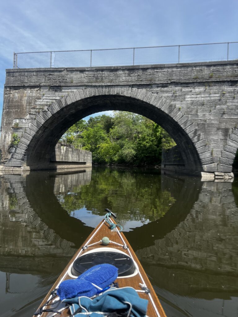 The tip of a brown kayak pointed toward an ancient stone arch in a river. The arch is what is left of an aqueduct that carried the old Erie Canal over the Mohawk River.